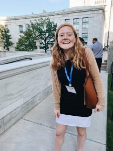 Smiling young woman with long red hair wearing a black dress and cardigan, standing outdoors near a marble building.