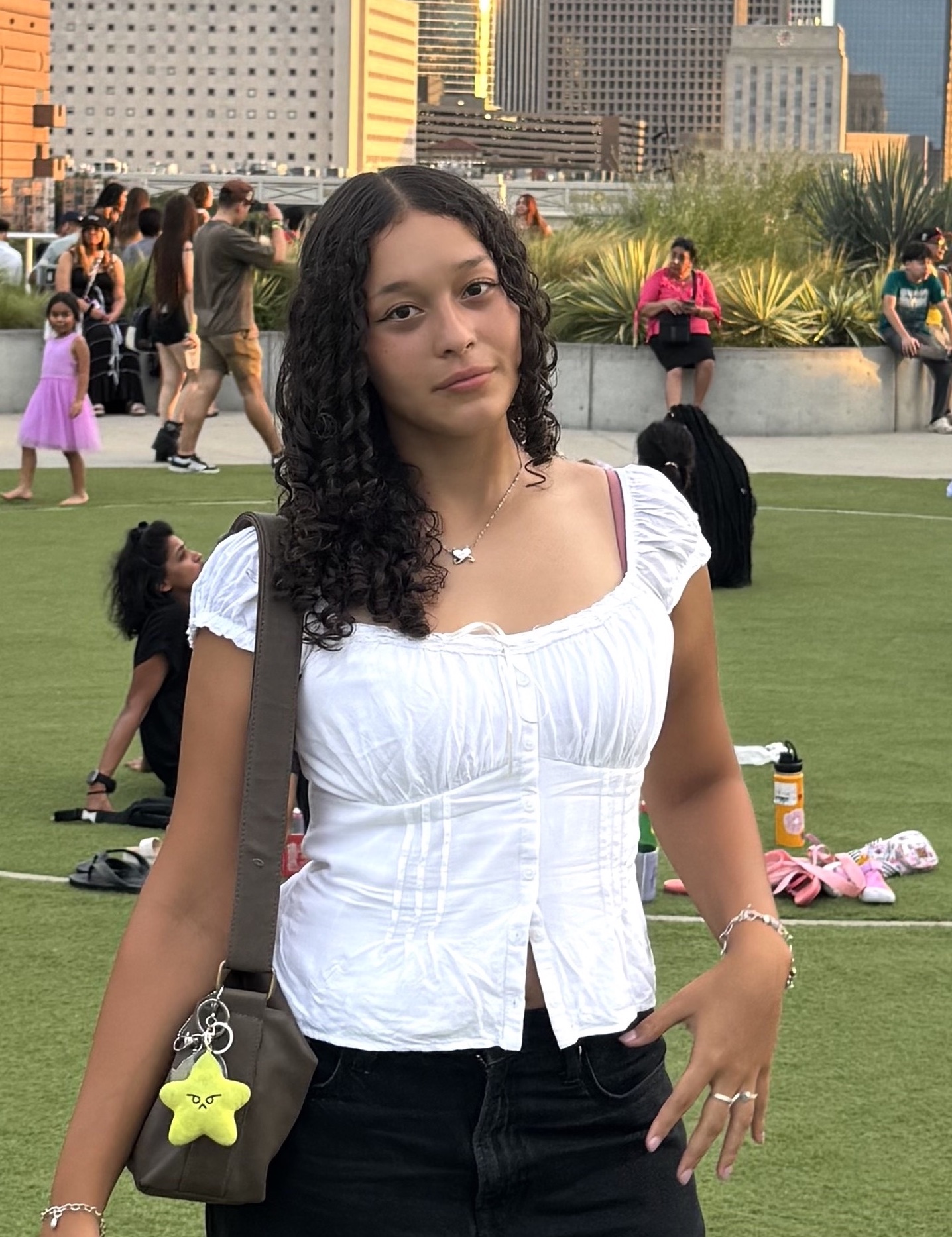 Young woman with curly hair wearing a white top and black pants, standing on green grass with people in the background.