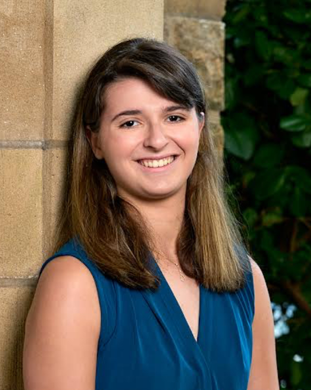 Smiling young woman in a blue top standing against a stone wall with greenery in the background.