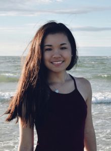 Young woman with long hair smiles at the beach, standing in front of ocean waves under a partly cloudy sky.