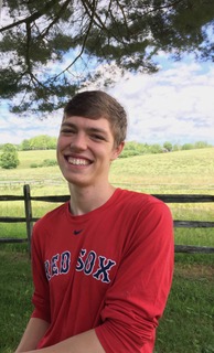 Smiling young man in a red shirt stands outdoors with a green landscape and blue sky in the background.