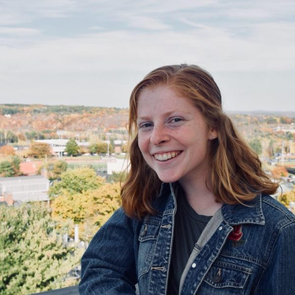 Young woman with red hair smiles while standing outdoors with a scenic view of trees and buildings in the background.