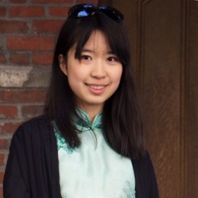 Young woman wearing a green top and black outerwear, smiling outdoors with brick wall in the background.