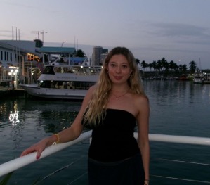 Woman in a strapless top stands near a waterfront at dusk, with a boat and palm trees in the background.