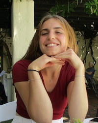 Young woman smiling with her hands under her chin, sitting outdoors with greenery and people in the background.